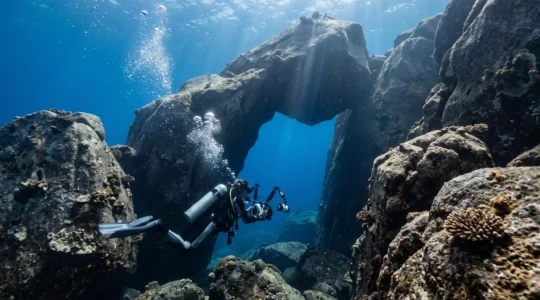 A scuba diver with an underwater camera rig swims toward a massive granite boulder archway in the clear blue water of the Similan Islands.