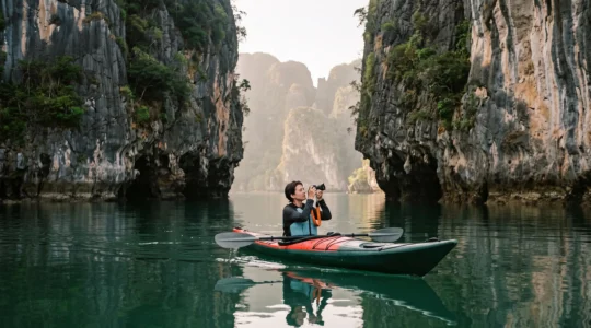 A photographer in a sea kayak paddles at water level between the sheer limestone karst towers of Phang Nga Bay, composing a shot of the cliffs reflected in the calm early-morning water.