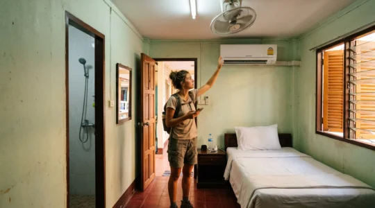 A traveller inspects the wall-mounted air-conditioning unit in a simple Thai guesthouse room with a ceiling fan and open bathroom.